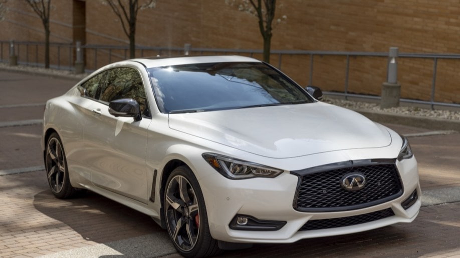 The 2022 Infiniti Q60 sits parked in front of a brick building. The car is white. We see it from a front quarter angle, facing to our right. It is sleek, with bulges along the sides of the hood and a long, sloping rear roofline.