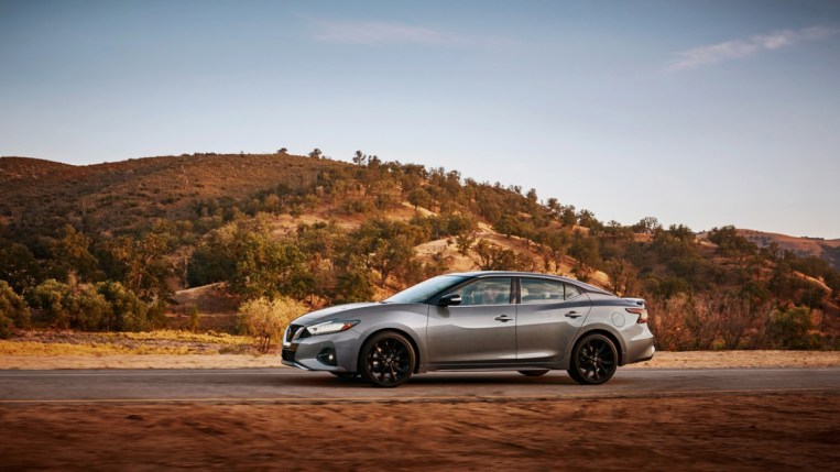 A 2022 Nissan Maxima drives past rolling hills. The car is grey with a black contrast-color roof. We see it in profile, facing to our left.