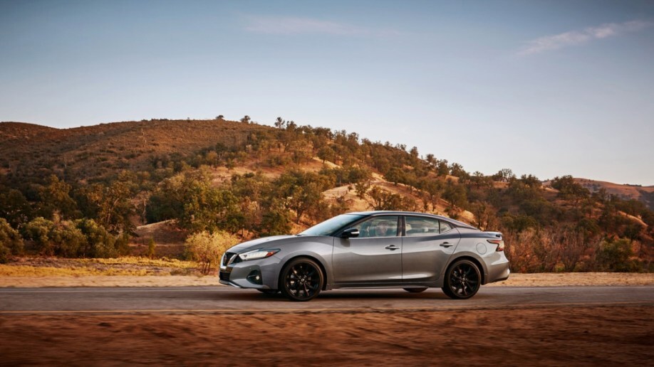 A 2022 Nissan Maxima drives past rolling hills. The car is grey with a black contrast-color roof. We see it in profile, facing to our left.