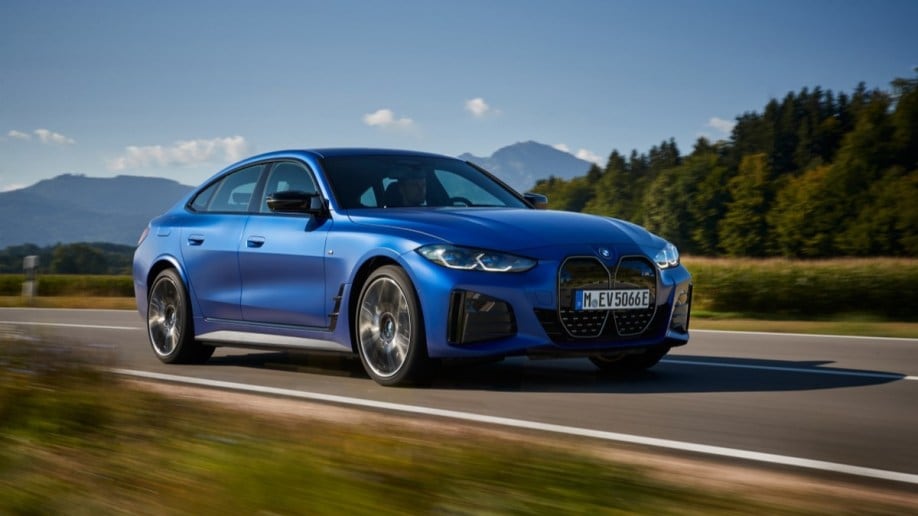 The 2023 BMW i4 electric sedan drives past a forest. The car is medium blue. We see it from a front quarter angle, facing slightly to our right. 