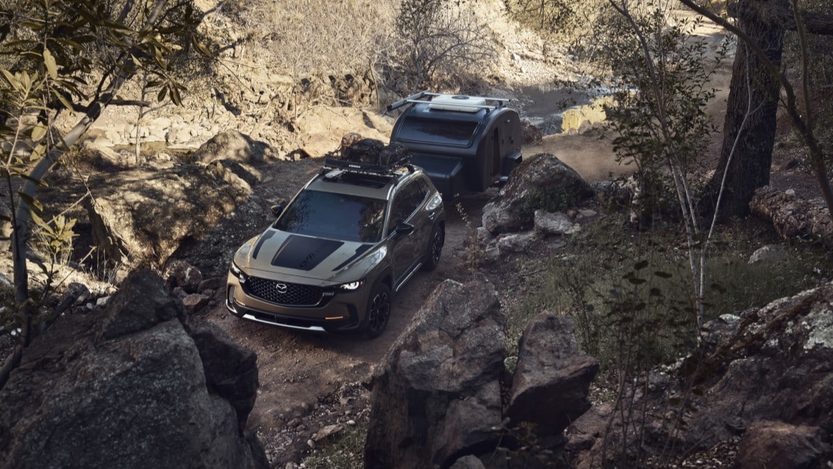 The 2023 Mazda CX-50 Meridian Edition tows a trailer down a forest trail. We see the SUV from overhead. It is a muted olive color Mazda calls Zircon Sand, with matte black hood decals and a loaded roof rack.