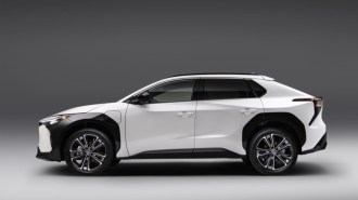 The 2023 Toyota bZ4X sits parked against a gray wall. The car is white, with thick black body cladding around the front wheels. We see it in profile.