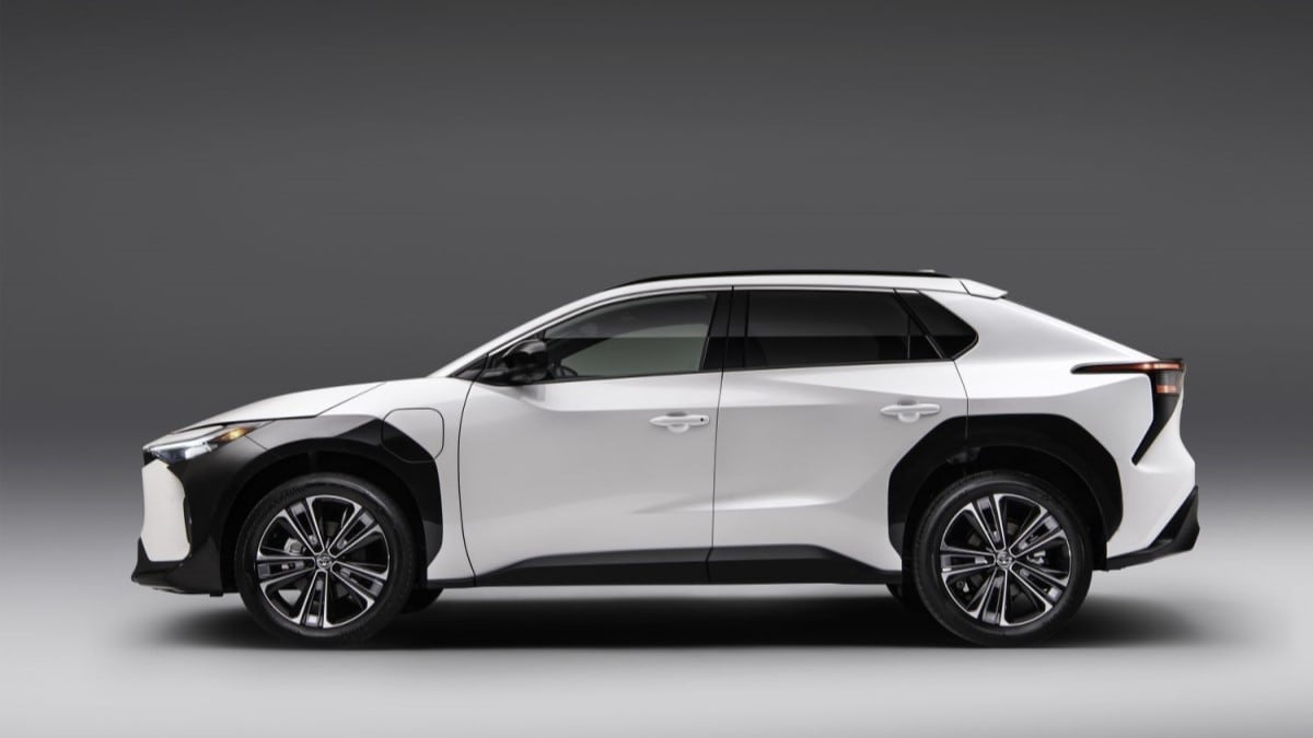 The 2023 Toyota bZ4X sits parked against a gray wall. The car is white, with thick black body cladding around the front wheels. We see it in profile.
