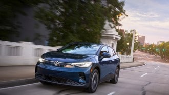 The 2023 Volkswagen ID.4 drives down a city street. The crossover is medium blue. We see it from a front quarter angle, facing slightly to our left.