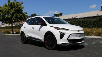 White 2023 Chevrolet Bolt EUV Premier near a tree and green hedge.