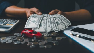 A pair of hands holding hundred dollar bills next to a toy car