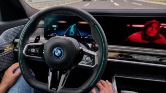 A driver demonstrates BMW's Personal Pilot L3 system with a video playing on the car's screen and the driver's hands off the steering wheel