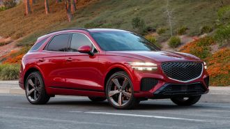 2024 Genesis GV70 in red with shrubs and trees in the background.