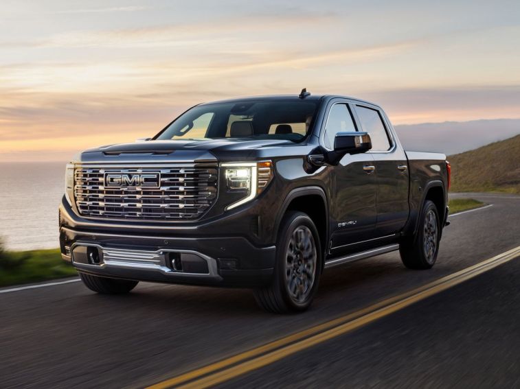 2024 GMC Sierra 1500 Denali Ultimate in Titanium Rush Metallic driving on a road next to water.