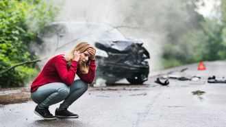 A young woman with smartphone by the damaged car after a car accident, making a phone call.